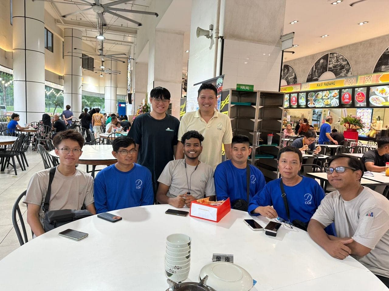 Members of the COF team, several in COF-branded shirts, smiling around a round table at a food court during the annual dinner.