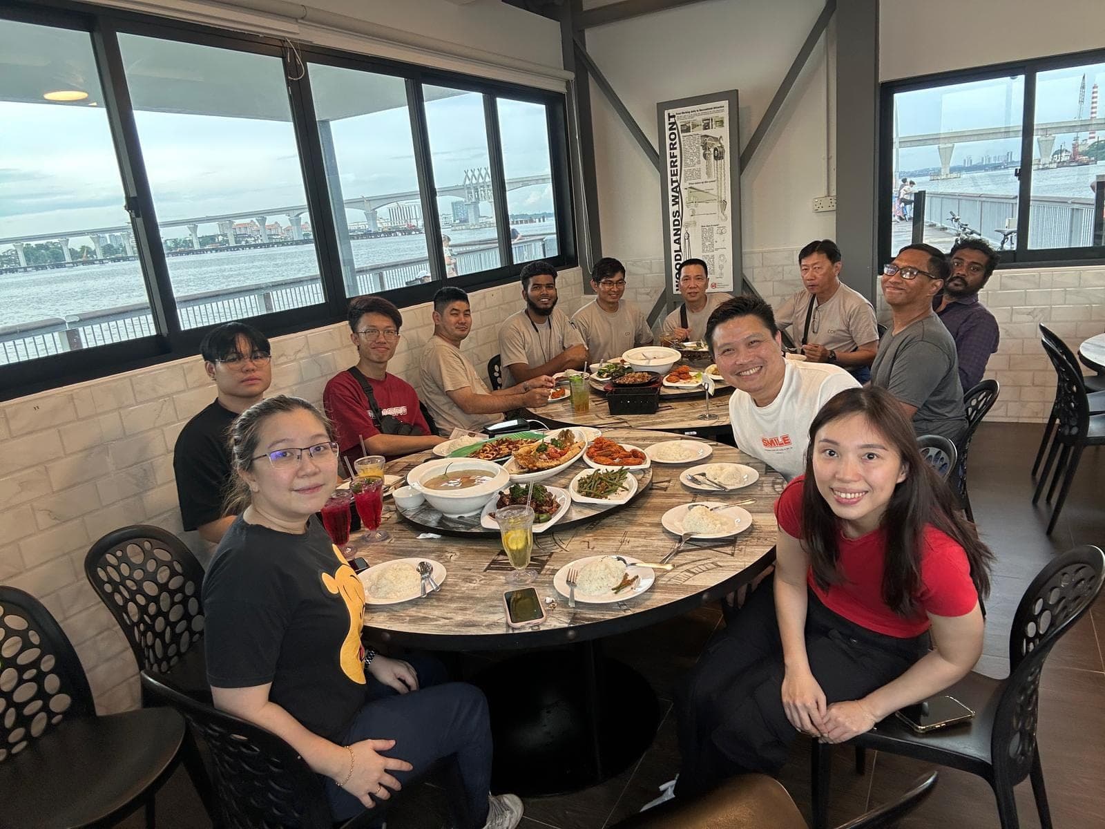 COF staff seated around a large round table of shared dishes at a waterfront restaurant, with the sea and a bridge visible through the windows.