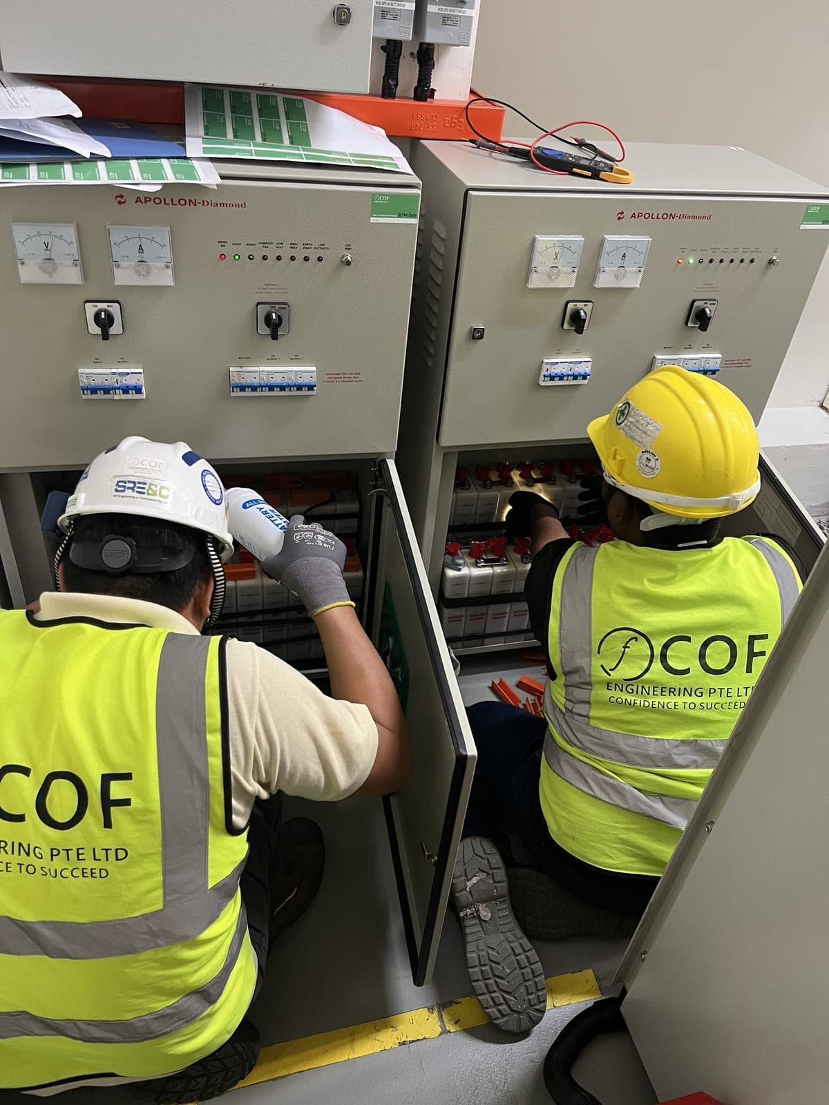 Two COF technicians kneel at open battery charger cabinets, one topping up battery cells, with a clamp meter resting on top of the cabinet.