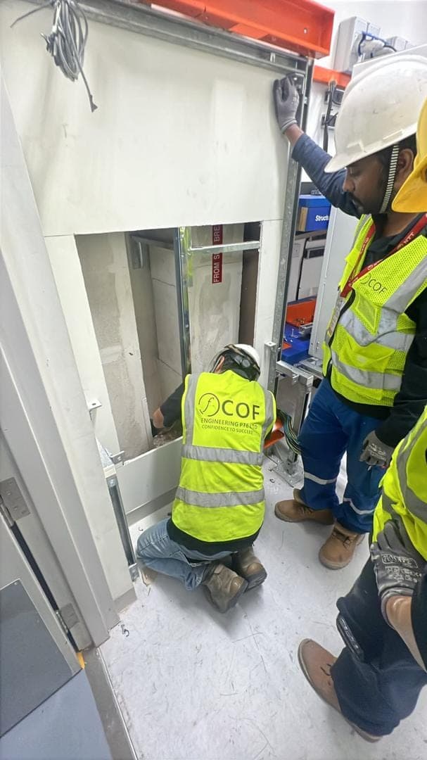 A COF technician kneels inside a cable riser opening while two colleagues stand by next to the cable tray.