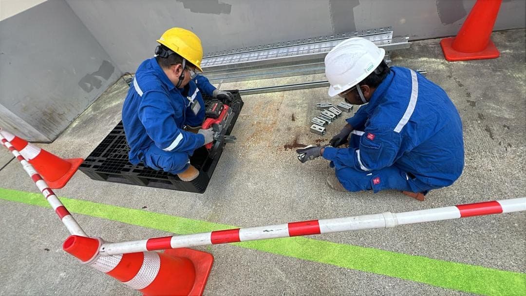 Two technicians in blue coveralls cut and fit cable tray brackets inside a coned-off work area.