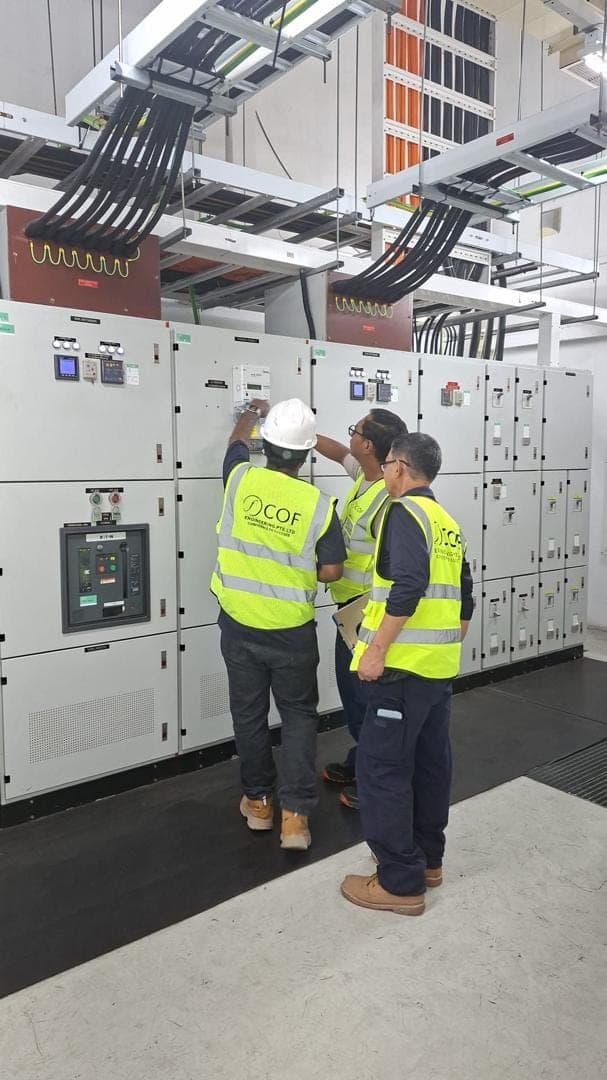 Three COF engineers in hi-vis vests reading a meter on an LV main switchboard, with cable ladders running overhead.