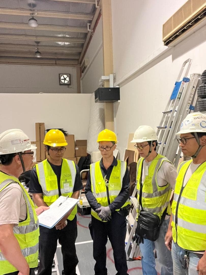 A supervisor with a clipboard briefs five COF staff in helmets and vests before work starts.