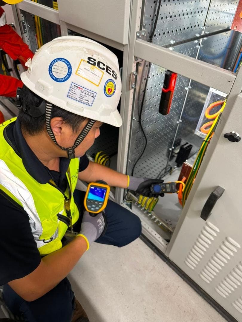 A COF engineer kneels at an open LV panel holding a thermal imager in one hand and a clamp meter on a cable in the other.
