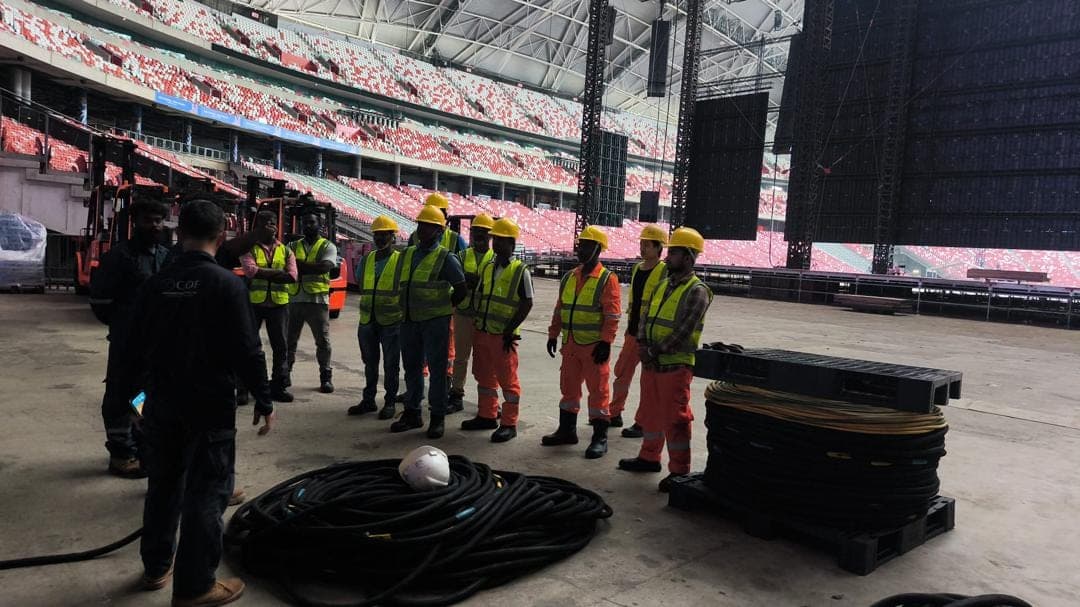 A crew of about a dozen workers in helmets and vests listens to a toolbox briefing beside coiled cables inside a large indoor venue.